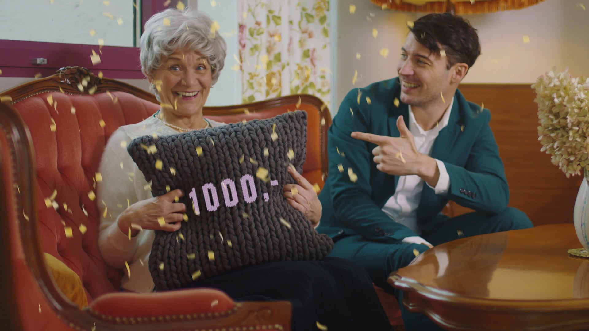 Ältere Frau hält ein Kissen mit der Aufschrift 1000 Euro, während ein Mann neben ihr auf dem Sofa sitzt und auf sie zeigt