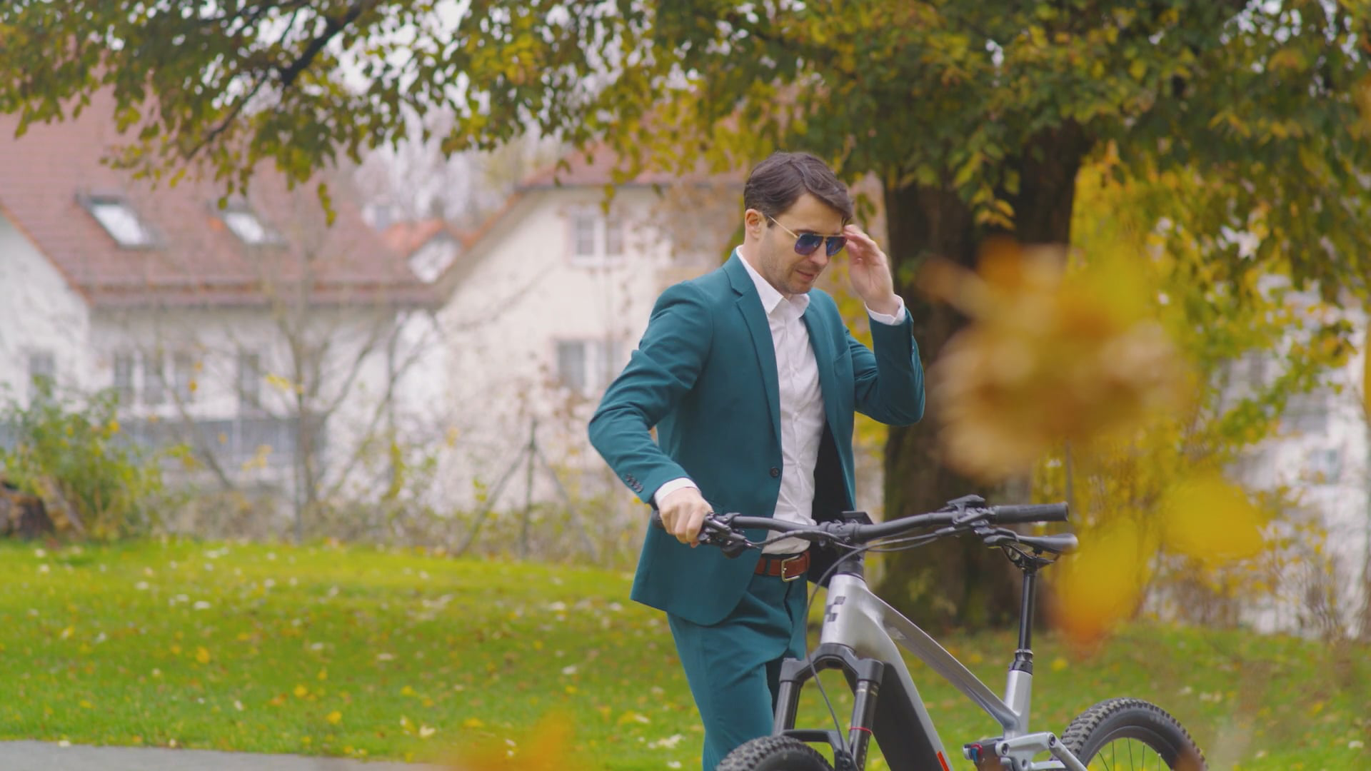 Mann im türkisfarbenen Anzug schiebt ein hochwertiges E-Mountainbike durch einen herbstlichen Park